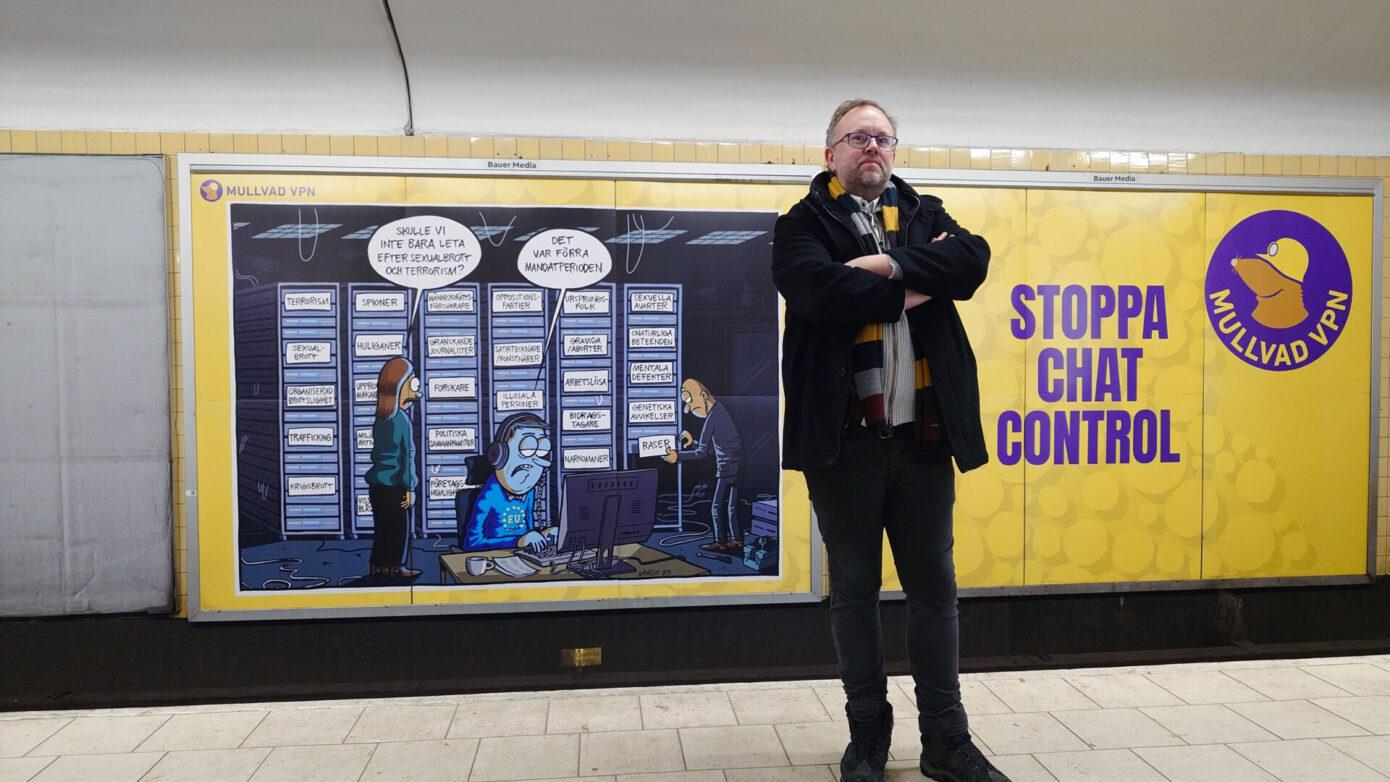 Mikael Enmalm står med armarna i kors i Stockholms tunnelbana framför en stor gul Mullvad-annons med budskapet: Stoppa Chat Control.