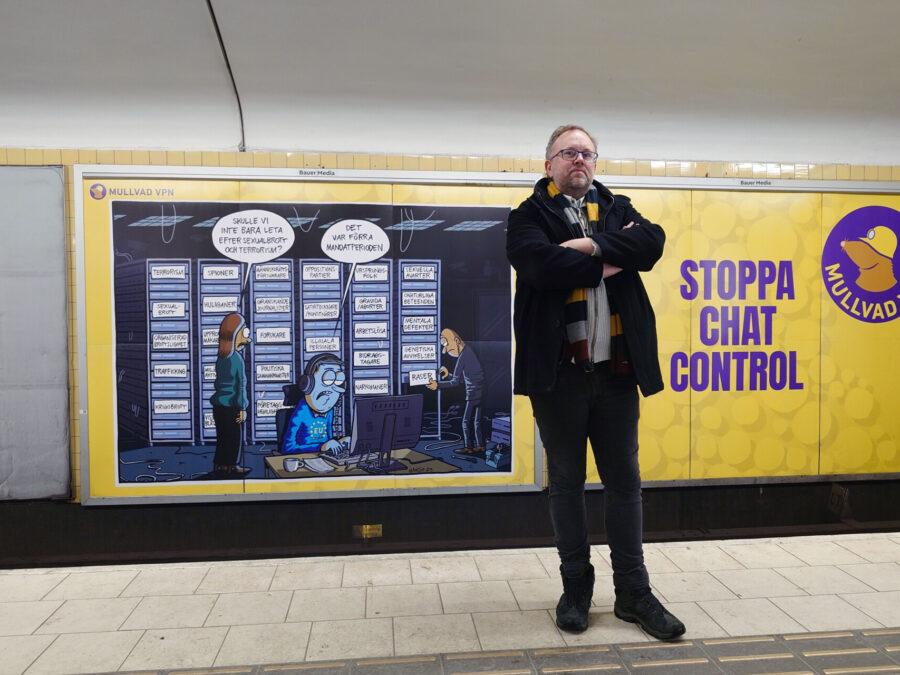 Mikael Enmalm står med armarna i kors i Stockholms tunnelbana framför en stor gul Mullvad-annons med budskapet: Stoppa Chat Control.